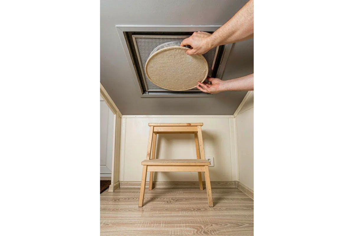 Technician replacing a round filter element in a ceiling vent of a prefab home from a step stool