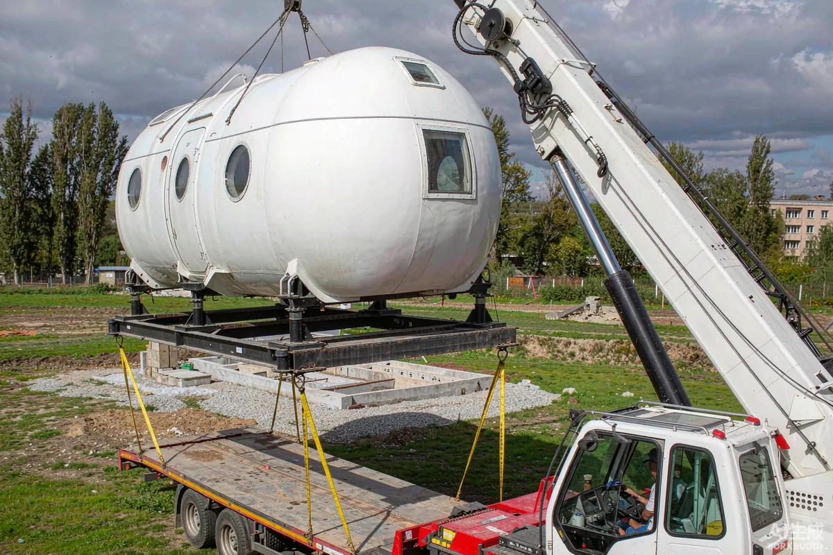 Prefab capsule home being lifted by a mobile crane off foundation blocks onto a flatbed trailer